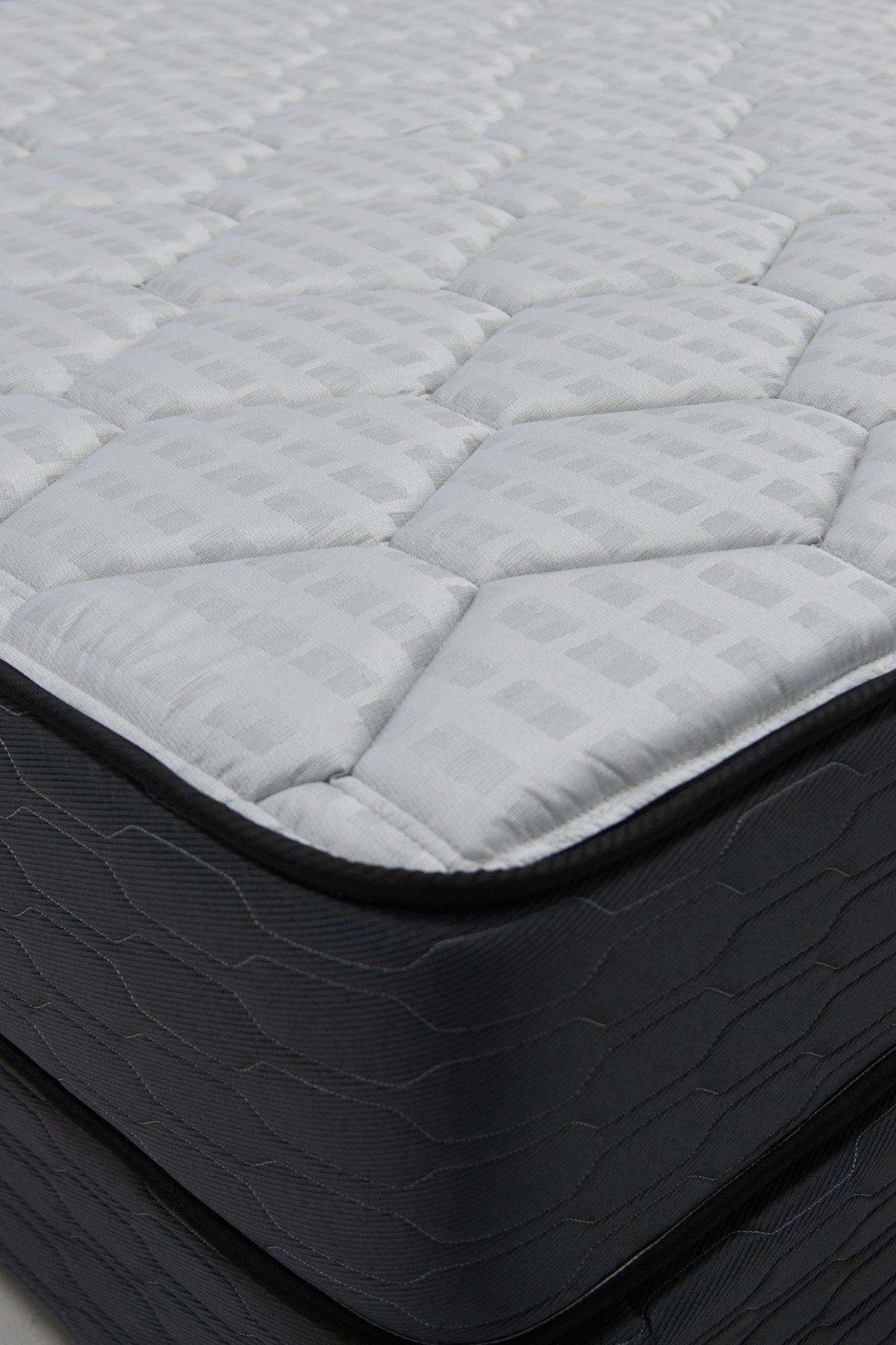Quina de um colchão sem roupa de cama, com a face pespontada, a lateral escura e a base box embaixo Quina de um colchão sem roupa de cama, com a face pespontada, a lateral escura e a base box embaixo